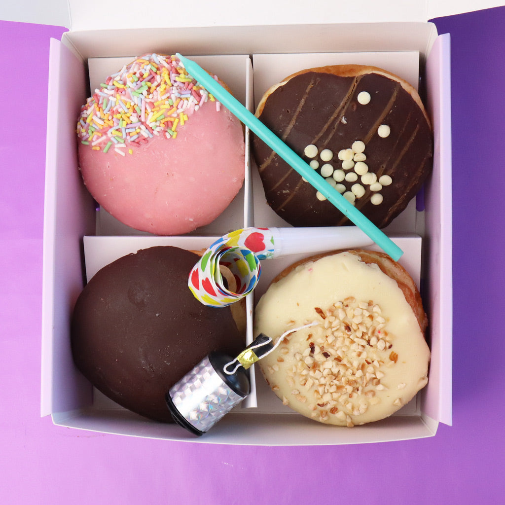 Box of assorted donuts with a candle, blower and popper on a purple background