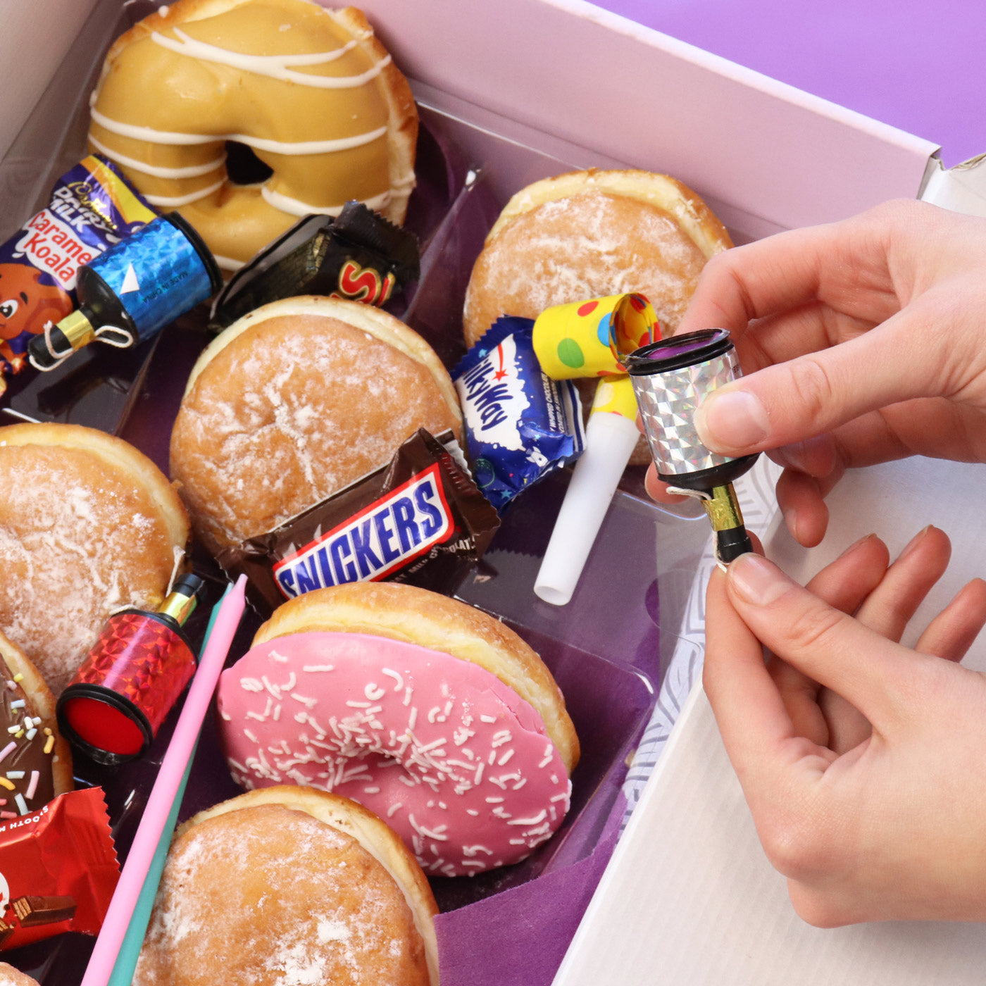 Box of assorted donuts with Snickers and other funsized chocolates, and a hand holding a birthday popper.
